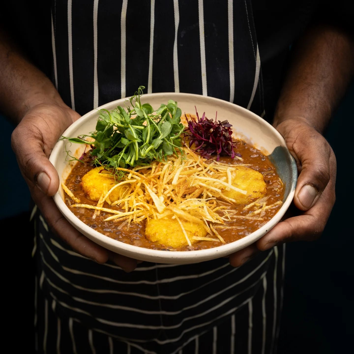 Chef Dhayalan's hands offering a bowl of curry dressed with pea shoots and crisps, striped apron behind